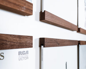 Close-up of a wall-mounted framed vinyl record display with walnut hardwood floating shelf that line the top and bottom on the vinyl record holding them against a white wall. 