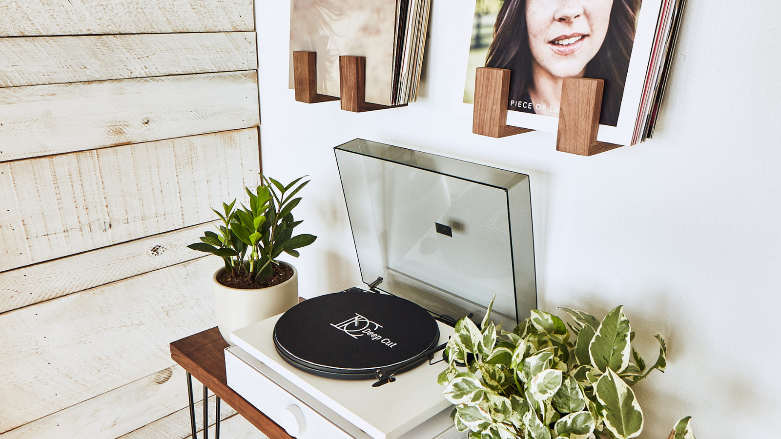 A white Andover SpinDeck turntable sitting on a white SpinBase speaker system on a walnut table with plants on both side and wall mounted front facing record shelves above